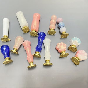 Könnte beinhalten: Eine Sammlung von 13 bunten Wachssiegel-Stempeln mit verschiedenen Designs, darunter Pfotenabdrücke, eine Katze und eine Perlenkette. Die Stempel sind aus Harz und haben eine goldfarbene Basis.