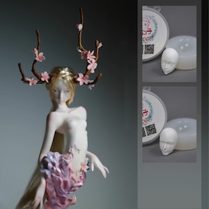 May include: A white porcelain doll with long blonde hair and a delicate face. The doll is wearing a pink and white floral crown and a pink and white floral skirt. The doll is also wearing a pair of white wings. The doll is standing on a white base. The image also shows two silicone moulds for making doll heads.