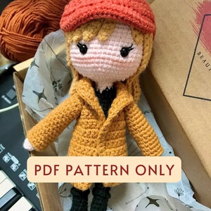 Könnte beinhalten: Eine gehäkelte Puppe mit roter Mütze, gelbem Mantel und schwarzen Stiefeln. Die Puppe sitzt in einer Schachtel mit Seidenpapier. Der Text "PDF PATTERN ONLY" befindet sich auf einem rot-orangenen Banner.