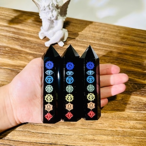 Könnte beinhalten: Drei schwarze Obsidian-Kristallstäbe mit Chakra-Symbolen in verschiedenen Farben. Die Stäbe werden in einer Hand gehalten, mit einer kleinen weißen Engelsstatue im Hintergrund. Die Stäbe sind poliert und haben eine spitze Spitze.