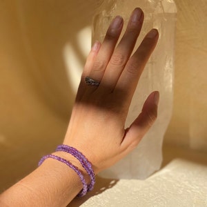 Peut inclure: Une main portant une bague en argent et une pile de trois bracelets en améthyste, touchant un grand cristal de quartz clair.