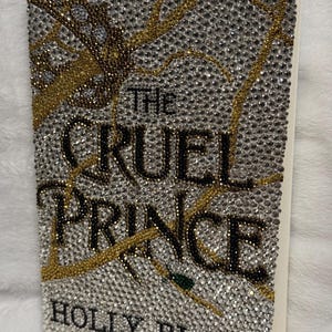 Pu&ograve; includere: Una copertina di libro con un design scintillante che presenta un albero con strass dorati e argentati. Il titolo "The Cruel Prince" &egrave; scritto in lettere nere. L'autore "Holly Black" &egrave; scritto in lettere nere nella parte inferiore della copertina.