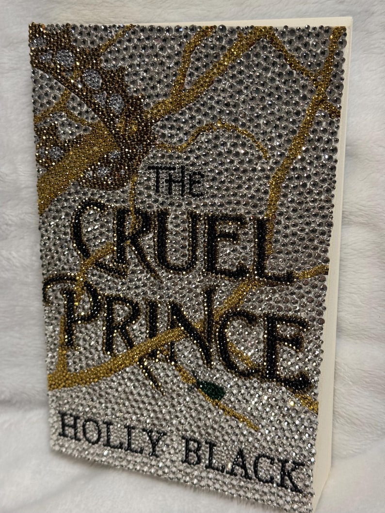 Pu&ograve; includere: Una copertina di libro con un design scintillante che presenta un albero con strass dorati e argentati. Il titolo "The Cruel Prince" &egrave; scritto in lettere nere. L'autore "Holly Black" &egrave; scritto in lettere nere nella parte inferiore della copertina.