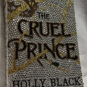 Pu&ograve; includere: Una copertina di libro con il titolo "The Cruel Prince" di Holly Black. La copertina &egrave; ricoperta di strass, con un design oro e argento.