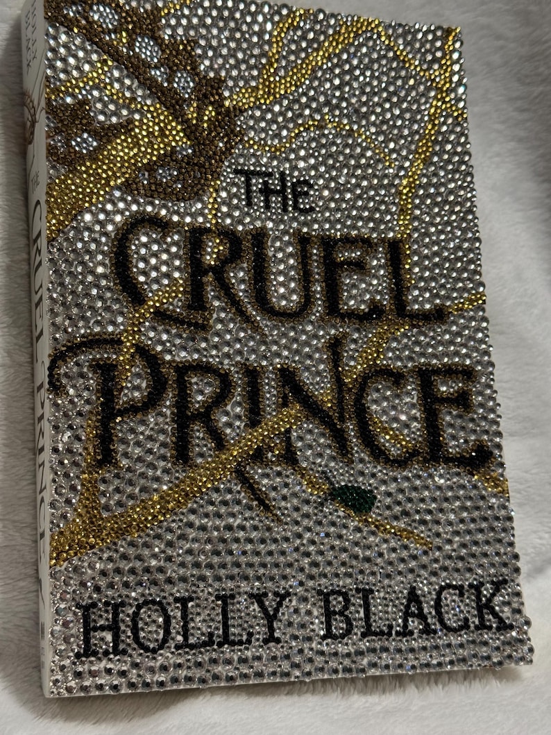 Pu&ograve; includere: Una copertina di libro con il titolo "The Cruel Prince" di Holly Black. La copertina &egrave; ricoperta di strass, con un design oro e argento.