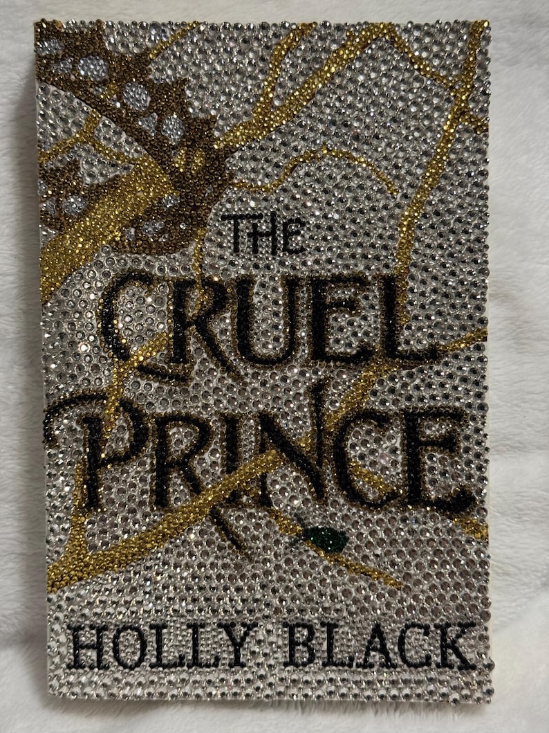 Pu&ograve; includere: Una copertina di libro con un design argento, oro e nero. Il titolo "The Cruel Prince" &egrave; scritto in lettere nere. L'autore "Holly Black" &egrave; scritto in lettere nere nella parte inferiore della copertina.