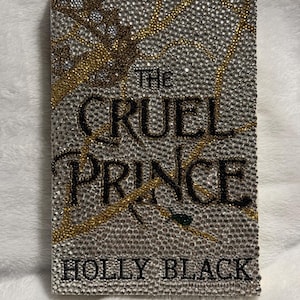 Pu&ograve; includere: Una copertina di libro con sfondo argento e oro ricoperto di strass. Il titolo "The Cruel Prince" &egrave; in lettere nere. L'autore "Holly Black" &egrave; in lettere nere nella parte inferiore della copertina.