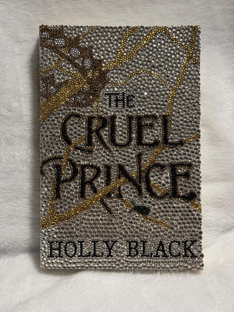 Pu&ograve; includere: Una copertina di libro con sfondo argento e oro ricoperto di strass. Il titolo "The Cruel Prince" &egrave; in lettere nere. L'autore "Holly Black" &egrave; in lettere nere nella parte inferiore della copertina.