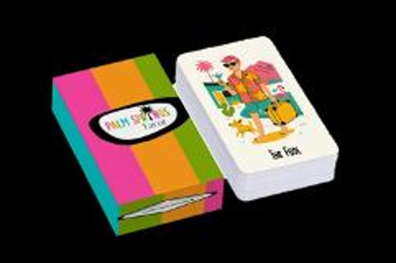 Peut inclure: Un jeu de cartes et son &eacute;tui. La bo&icirc;te pr&eacute;sente des rayures verticales roses, oranges, vertes et jaunes. Le texte "Palm Springs" est dans un ovale noir. Les cartes montrent un personnage de dessin anim&eacute; avec un chien, des palmiers et le texte "Eat Fast".