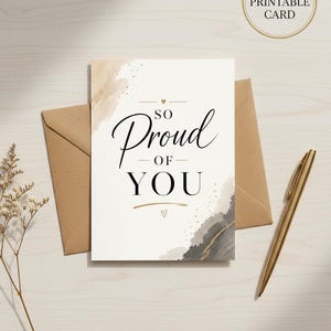 以下が含まれることがあります： SO PROUD OF YOUという文字が黒の筆記体とセリフ体で書かれた長方形のグリーティングカード。カードにはベージュとダークグレーの水彩画風の筆致とゴールドのアクセント、小さなハートのアイコンが描かれている。カードの後ろには茶色の封筒が置かれている。