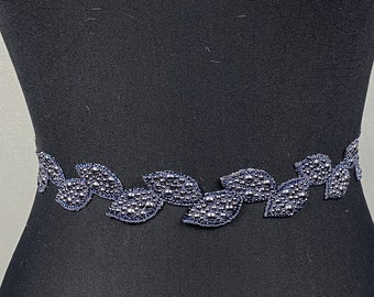May include: A dark gray beaded sash with a leaf design. The sash is made of small, round beads that are sewn together in a continuous pattern.