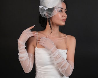 May include: A white, sheer, long-sleeved glove with a ruffled texture. The glove is worn on the left hand of a person wearing a white dress.
