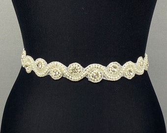 May include: A white beaded and rhinestone bridal belt with a wavy design. The belt is made of a delicate fabric and features a row of pearls and rhinestones.