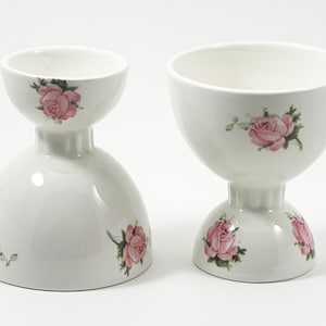 May include: Two white ceramic egg cups with pink rose designs. The egg cups have a wide, shallow top and a narrow base.