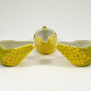 May include: A three-piece ceramic corn-shaped creamer and sugar bowl set. The set features a yellow and orange corn design with green husks and handles. The creamer and sugar bowl are on a white background.