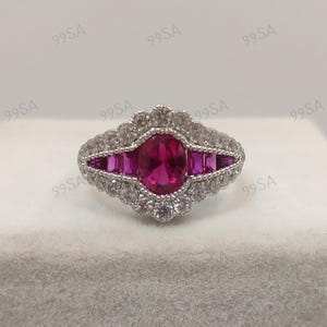 Könnte beinhalten: Ein silberner Ring mit einem großen ovalen Rubin-Mittelstein, flankiert von kleineren rechteckigen Rubinen. Der Ring ist von kleinen, klaren Akzentsteinen umgeben, die ein Design im Vintage-Stil schaffen. Der Ring liegt auf einer weißen Oberfläche.