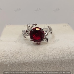 May include: A silver ring featuring a large, round, deep red gemstone. The ring's band is adorned with delicate, leaf-like designs, creating a nature-inspired aesthetic. The gemstone is the focal point, set prominently in the center.