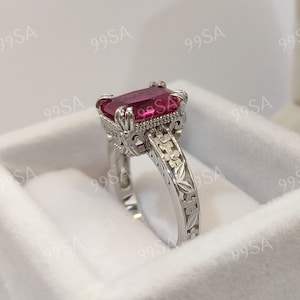 Peut inclure: Une bague en argent avec une grande pierre précieuse rouge de taille émeraude. La bande est décorée de motifs floraux.