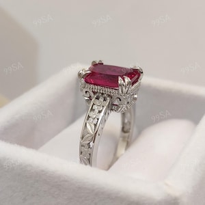 May include: A silver ring with a large, rectangular, red gemstone. The ring has an ornate, floral design on the band and around the gemstone setting. The ring is displayed in a white box.
