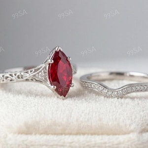 May include: A silver-toned ring set featuring a marquise-cut deep red gemstone centre stone with intricate filigree metalwork on the band, accompanied by a matching curved wedding band encrusted with small clear stones along the top edge.
