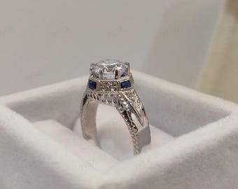 May include: A silver engagement ring with a large, clear center stone and two small, square blue stones. The ring has intricate filigree detailing on the band and is set in a white velvet box.