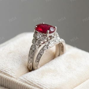 May include: A silver-coloured ring set featuring a large, oval-cut vibrant red gemstone as the centre stone. The band and the accompanying contoured band are crafted with intricate, engraved leaf patterns and small, round, clear accent stones.
