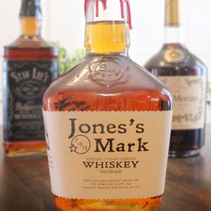 May include: A bottle of Jones's Mark Kentucky Straight Bourbon Whiskey with a red wax seal. The label reads "Handmade" and includes the distillery's information. Two other bottles are slightly out of focus in the background.