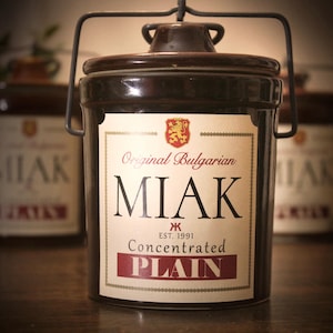 May include: A dark brown ceramic jar with a lid and a metal handle. The label reads "Original Bulgarian MIAK Concentrated PLAIN" with a crest and the text "EST. 1991". Other jars are visible in the background.