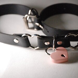 May include: A black leather choker with heart-shaped metal accents and a pink heart-shaped padlock.