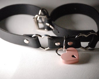 Könnte beinhalten: Ein schwarzes Lederhalsband mit herzförmigen Metallakzenten und einem rosafarbenen herzförmigen Vorhängeschloss.
