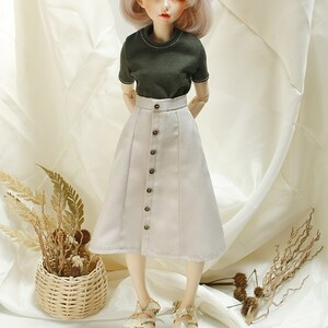May include: A doll wearing a green short-sleeved shirt and a beige skirt with buttons down the front. The doll has pink hair and blue eyes. The doll is wearing white sandals with brown straps.