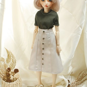 May include: A doll wearing a light brown skirt with a row of buttons down the front, a dark green short-sleeved shirt, and white sandals.