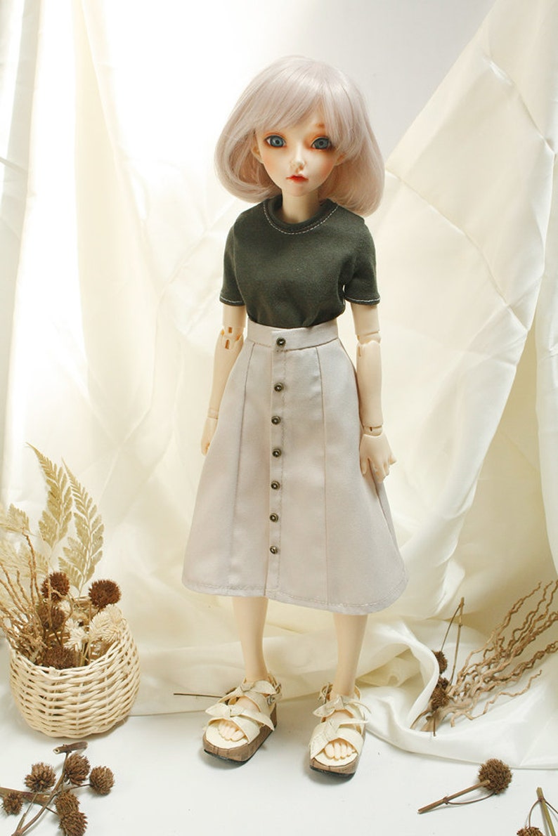 May include: A doll wearing a light brown skirt with a row of buttons down the front, a dark green short-sleeved shirt, and white sandals.