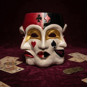 May include: A multi-faced mask with a harlequin design, featuring red, black, and white sections. Each face has a different expression and suit symbol. Playing cards and gold coins are scattered around the mask on a burgundy surface.