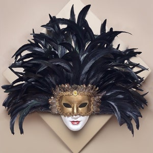May include: A black feathered masquerade mask with gold accents and a white face. The mask is decorated with feathers and has a gold and black design.