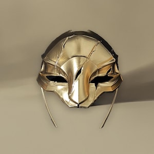 Pode incluir: Uma máscara dourada com um design estilizado. A máscara tem uma forma de rosto central com aberturas para os olhos e uma borda inferior pontiaguda. Duas finas peças de metal curvas se estendem dos lados. A máscara está em um fundo neutro.