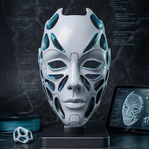 Op de afbeelding: Een wit masker met een futuristisch ontwerp, met open secties en blauwe accenten. Het masker wordt op een standaard getoond, met een klein model en een tablet met een 3D-weergave. Het thema suggereert een mix van kunst en technologie.