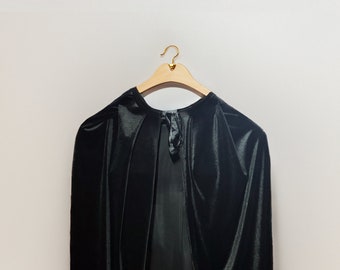 Op de afbeelding: Een zwarte fluwelen cape die aan een houten hanger met een gouden haak hangt.