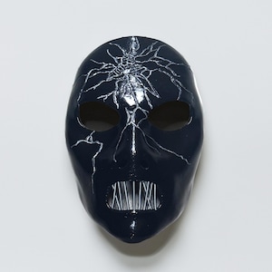 Puede incluir: Una máscara azul marino oscuro con un diseño agrietado en forma de telaraña plateada. La máscara tiene aberturas para los ojos y la boca, con una rejilla metálica en la boca. El diseño sugiere una pieza teatral o de disfraz.