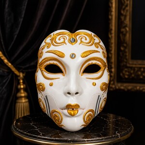 Könnte beinhalten: Eine weiße Vollgesichtsmaske mit aufwendigen, erhabenen goldenen Schnörkelmustern auf Stirn, Wangen und Kinn. Die Maske verfügt über mandelförmige Augenöffnungen, gold bemalte Lippen und zwei kleine silberfarbene runde Akzente auf der Stirn.