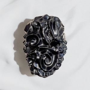 May include: A dark, textured, decorative object with an intricate, swirling design. The object has a metallic sheen and is set against a white background, creating a contrast that highlights its form and detail.