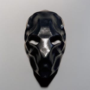 May include: A black mask with a sculpted, textured surface. The mask has eye openings and a contoured design, casting a shadow below. The mask appears to be made of a solid material.