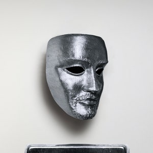 May include: A silver-colored mask with a human face, featuring a beard and mustache. The mask is made of a smooth, shiny material.