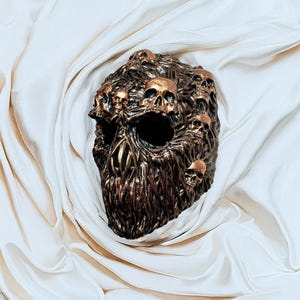 May include: A bronze-colored sculpture of a skull, intricately detailed with multiple smaller skulls emerging from its surface. The sculpture has a textured, organic appearance and is set against a backdrop of draped, off-white fabric.