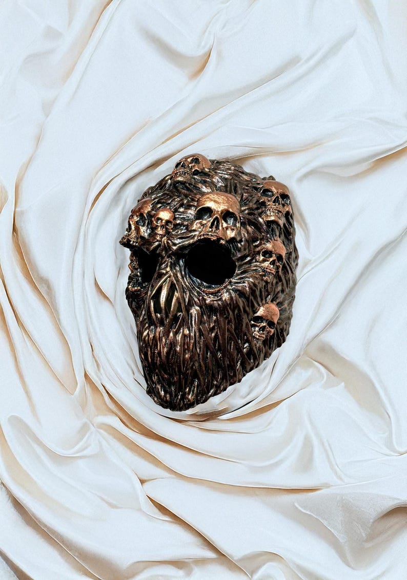 May include: A bronze-colored sculpture of a skull, intricately detailed with multiple smaller skulls emerging from its surface. The sculpture has a textured, organic appearance and is set against a backdrop of draped, off-white fabric.