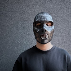 May include: A person wearing a black mask with a cracked design and a metal mouth guard. The mask has a white crackle design and a silver metal mouth guard.