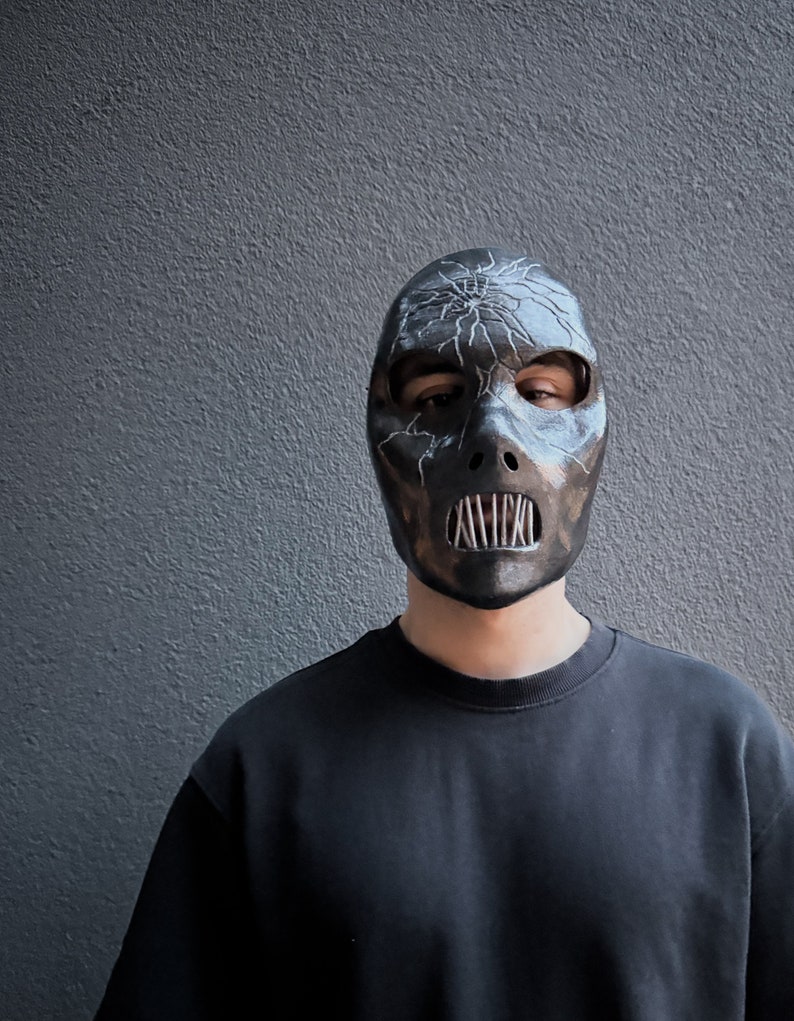 May include: A person wearing a black mask with a cracked design and a metal mouth guard. The mask has a white crackle design and a silver metal mouth guard.