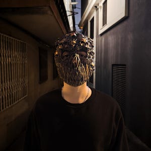 May include: A detailed, ornate mask with a dark, metallic finish. The mask features intricate skull-like details and a textured surface. The person is wearing a black long-sleeve shirt and a silver chain necklace. The background is a dark alley.