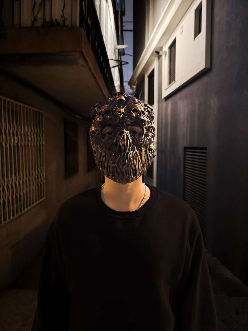 May include: A detailed, ornate mask with a dark, metallic finish. The mask features intricate skull-like details and a textured surface. The person is wearing a black long-sleeve shirt and a silver chain necklace. The background is a dark alley.