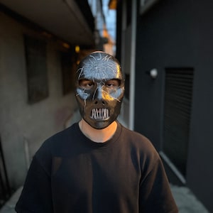 May include: A person wearing a black mask with a cracked design and a metal mouth guard. The mask is covering the person's face, and they are standing in a narrow alleyway.